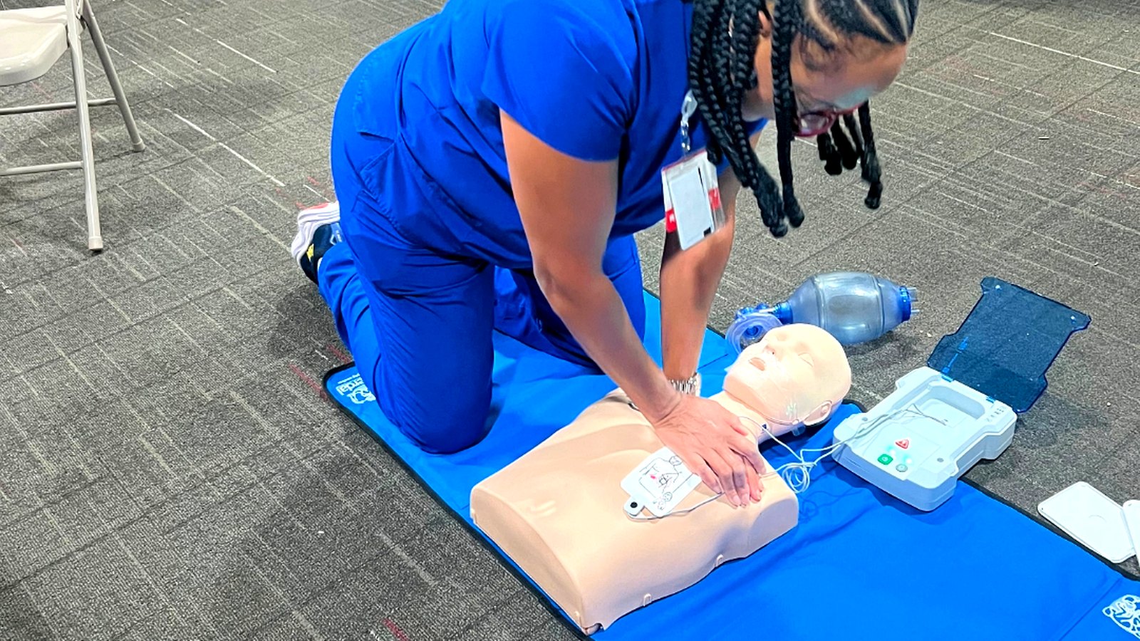 Nurse in blue scrubs performing chest compressions on an adult CPR manikin with an AED trainer beside her